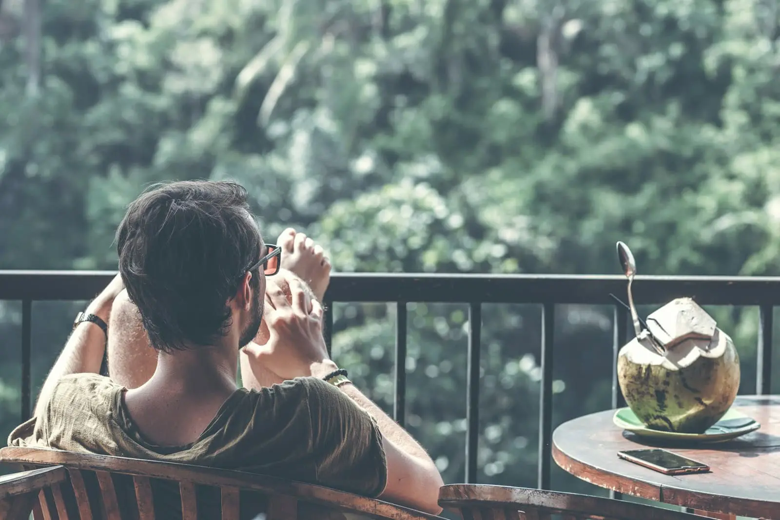 A man sits on a wooden chair, feet up on a balcony railing, looking at his phone. A table with a coconut drink and a smartphone is beside him, overlooking dense green foliage.