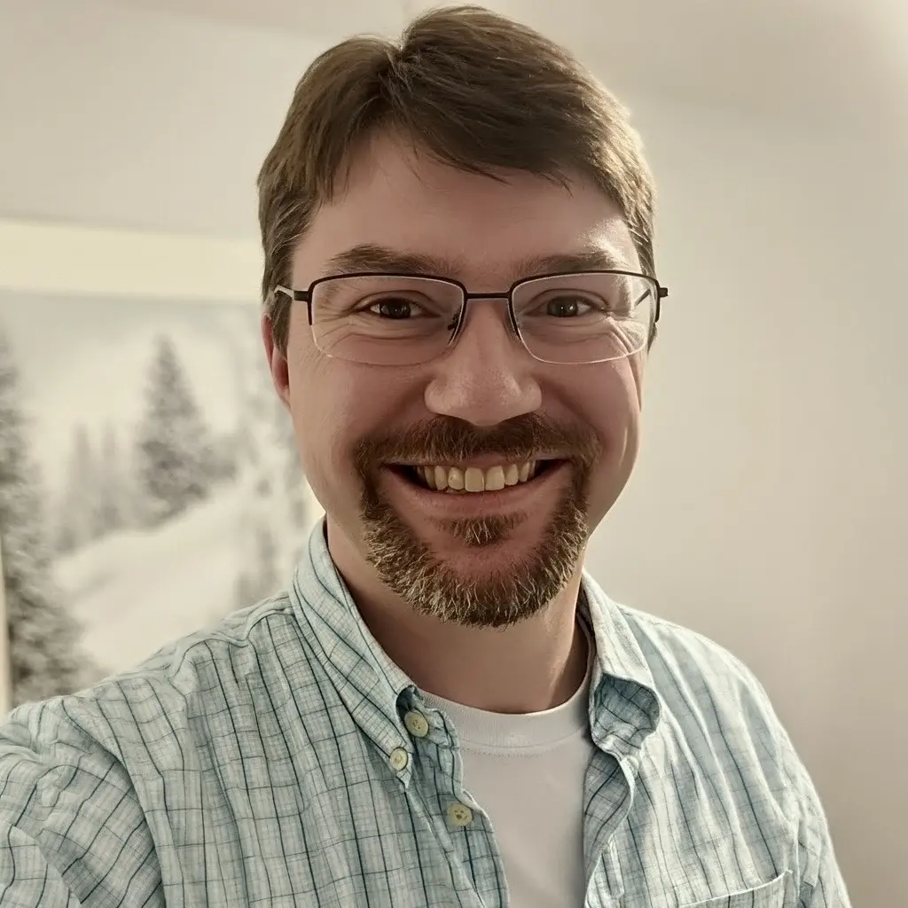 A man with glasses and a beard is smiling at the camera, wearing a light blue plaid shirt and a white t-shirt. A snowy landscape picture is visible in the background.