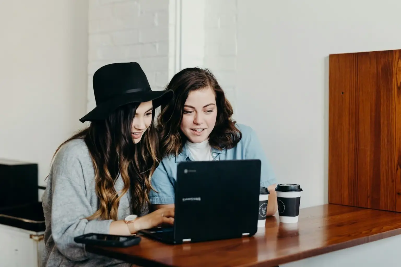 Two women sit at a wooden table, looking at a laptop screen together, with coffee cups beside them.